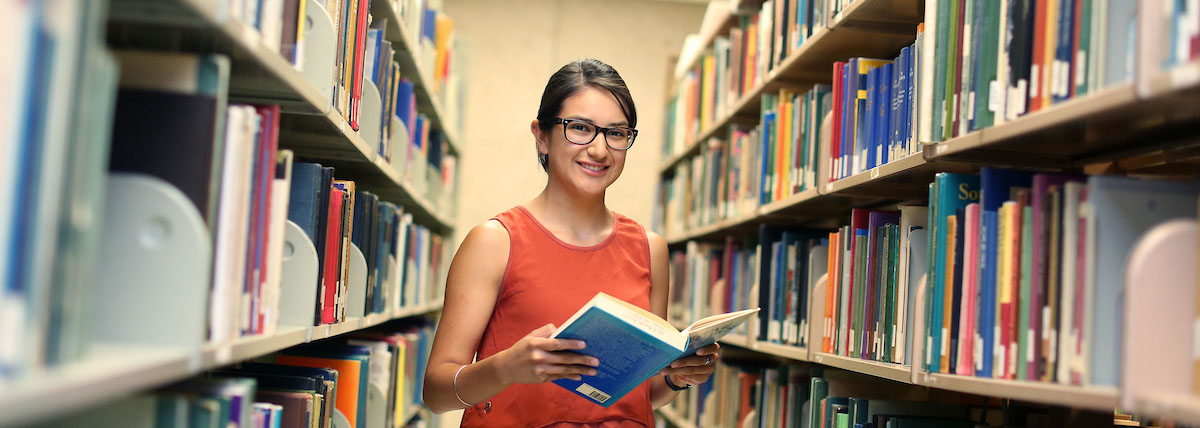 Image of a female student in the library between bookshelves holding a book and facing the camera. 