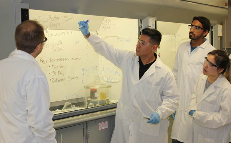 Dr Bachman listens to students wearing lab coats and safety glasses explain theory in lab