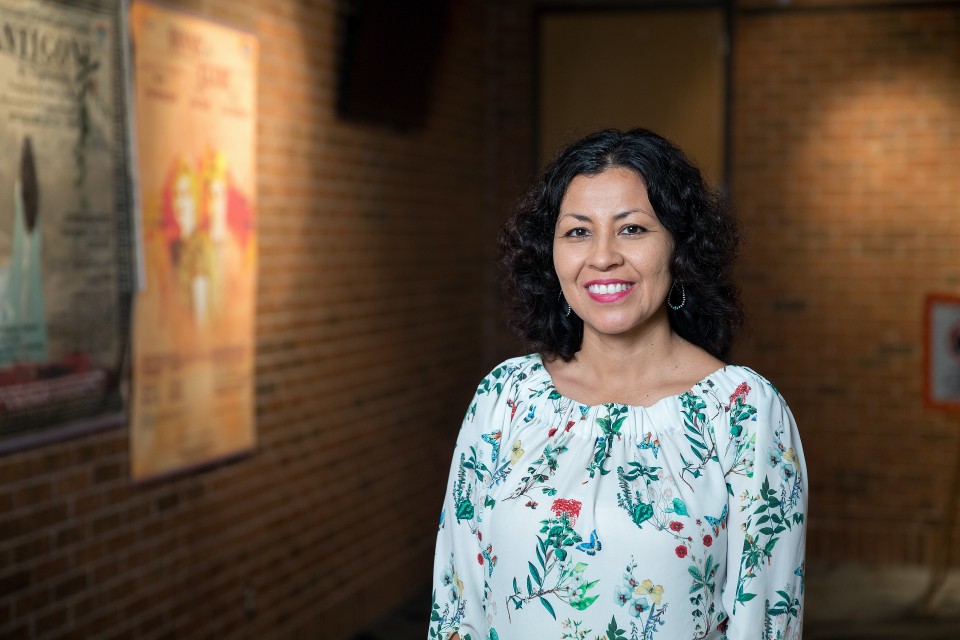 Photograph of a woman standing in a room, a wall behind her. 