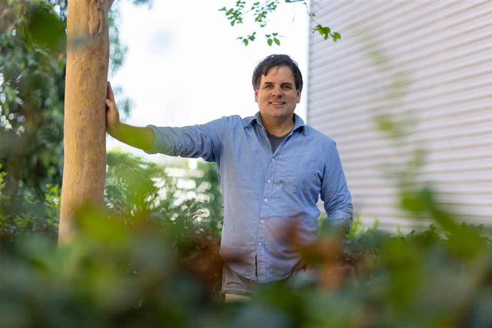 Faculty member stands outdoors on campus, leaning against a tree and smiling, with greenery in the foreground and a building in the background at Cal State LA.