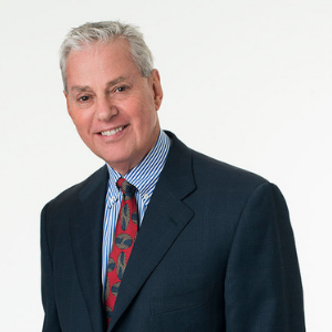 Man with gray hair, dark blue shuit, blue and white collared shirt, and red print tie