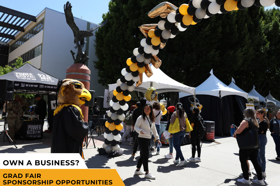 Eddie the Golden Eagle mascot dressed in Commencement regalia standing in front of a white, black, and gold balloon arch