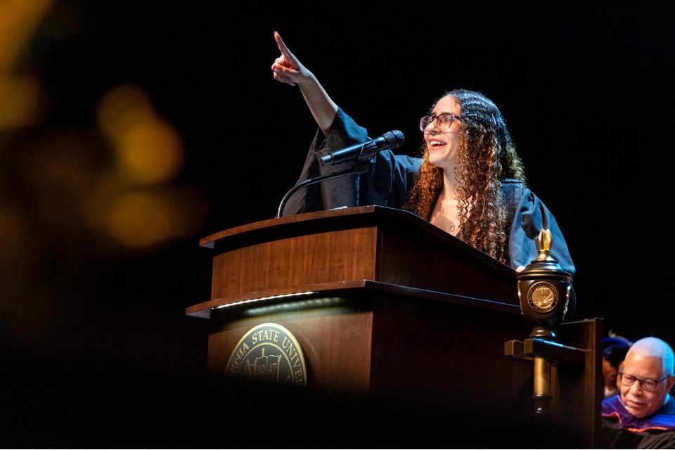 Cal State LA student delivers a commencement speech from a podium, pointing upward with enthusiasm while wearing a graduation gown.