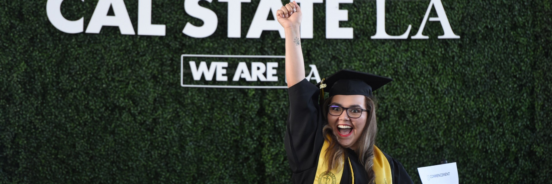 A woman in cap and gown smiles with her hand up