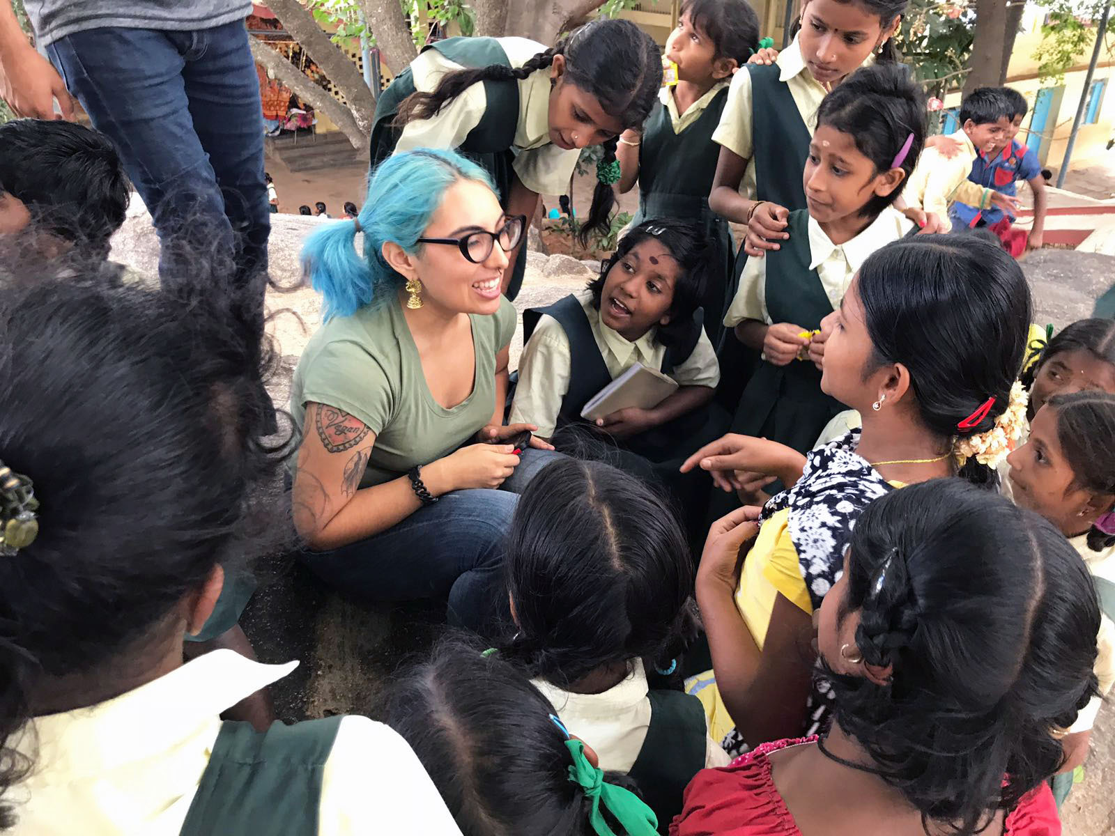 Smiling Cal State LA student sitting on the ground surrounded by listening young school students in India.