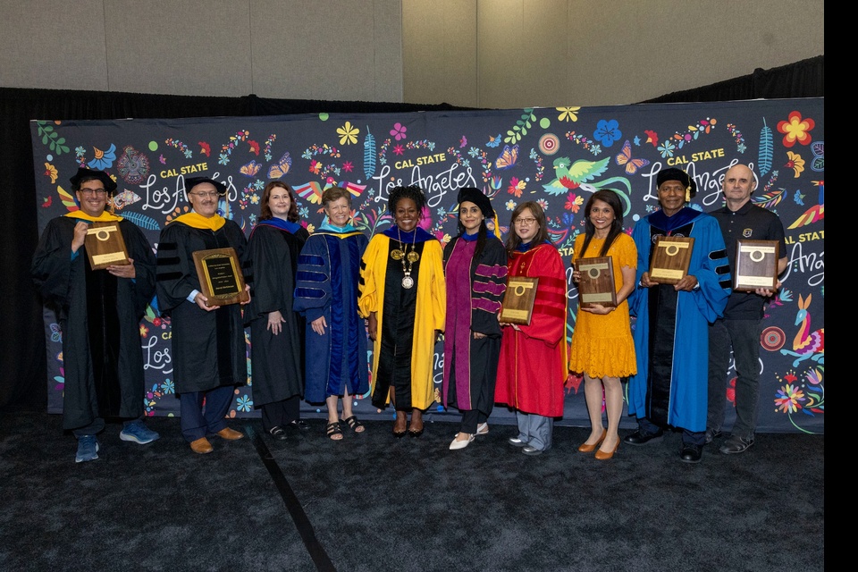 Cal State LA faculty and staff pose with plaques during an investiture ceremony, standing in front of a vibrant backdrop featuring floral and bird illustrations.