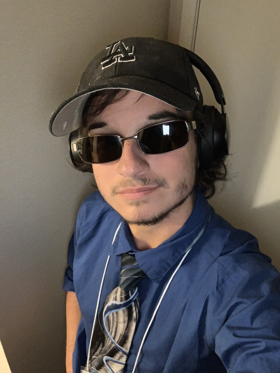 A photograph of Jayden Butler wearing a hat, glasses, and headphones wearing a blue shirt with a black, white, and blue tie against a cream background.