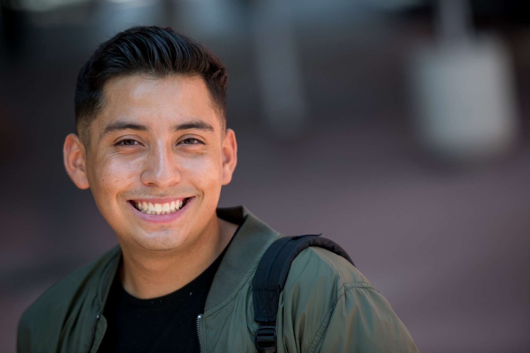 Cal State LA Downtown Student Marvin Vasquez smiles. He is wearing a dark green jacket and has a backpack on his arm.