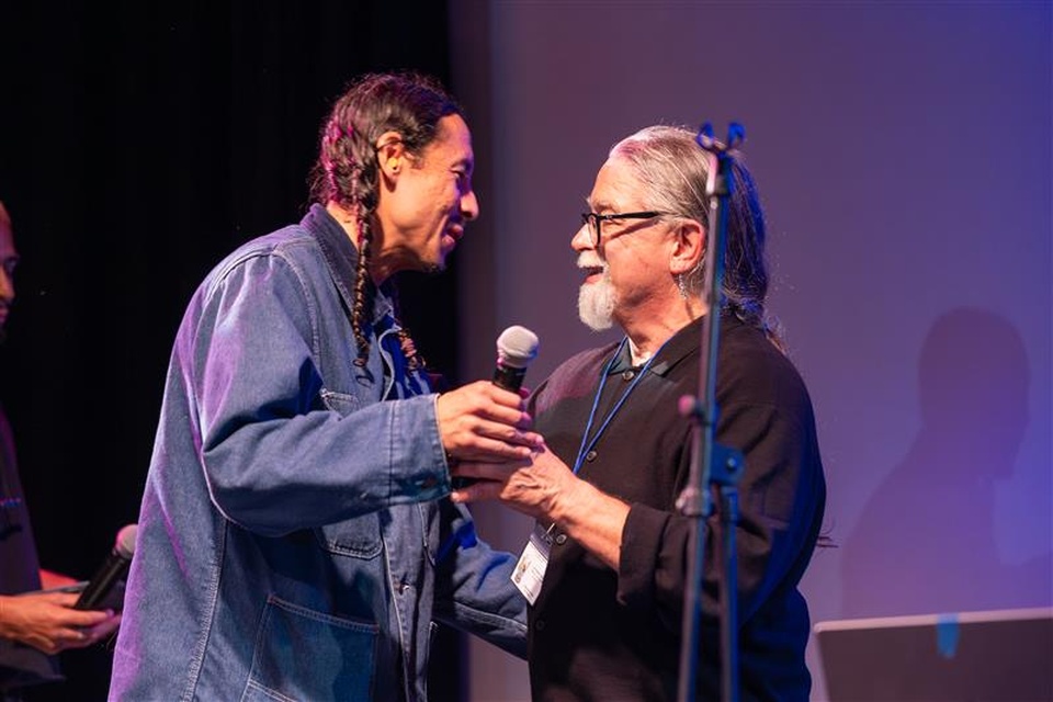 Two individuals greet and share a handshake on stage during the LAS 65 Years Celebration event at Cal State LA, under stage lighting with microphones visible.