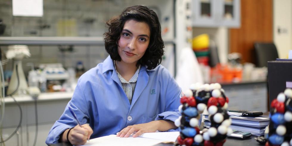 A student with short, dark hair wears a blue lab coat and holds a pipette in a lab