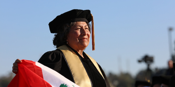 A doctoral student, Berenice Onofre-Vasquez, wears a gown, tam, and yellow stole and waves a Peruvian flag at commencement 2017