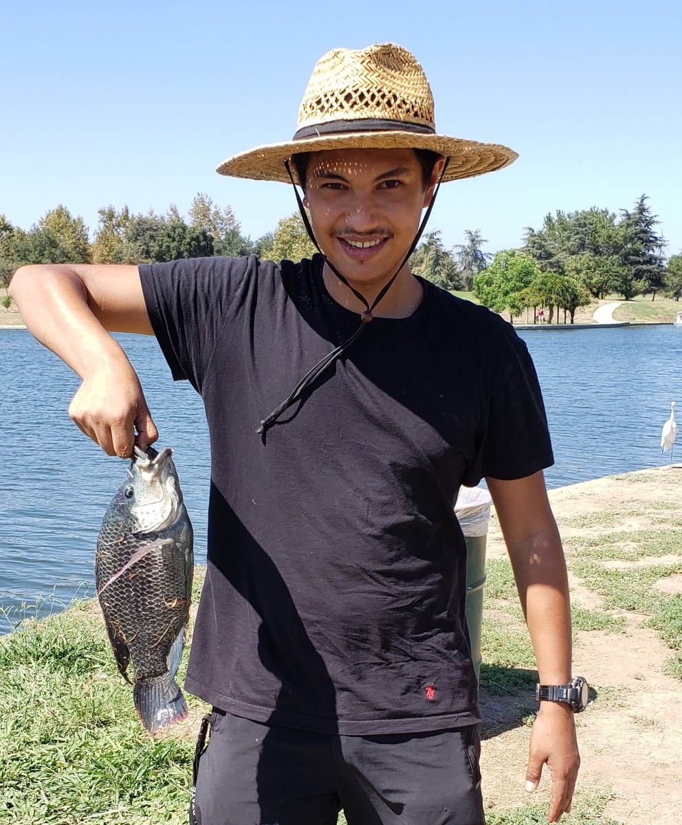 A photograph of Luis Valencia wearing a hat and a black shirt holding a fish by a lake.
