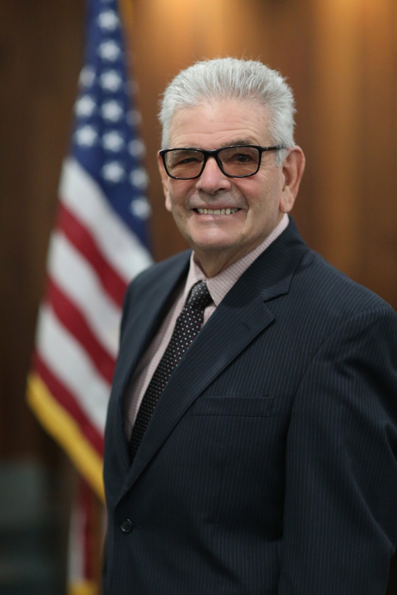 Headshot of man with gray hair and glasses in black suit, link pink shirt and black and white polka dot tie. US flag in background