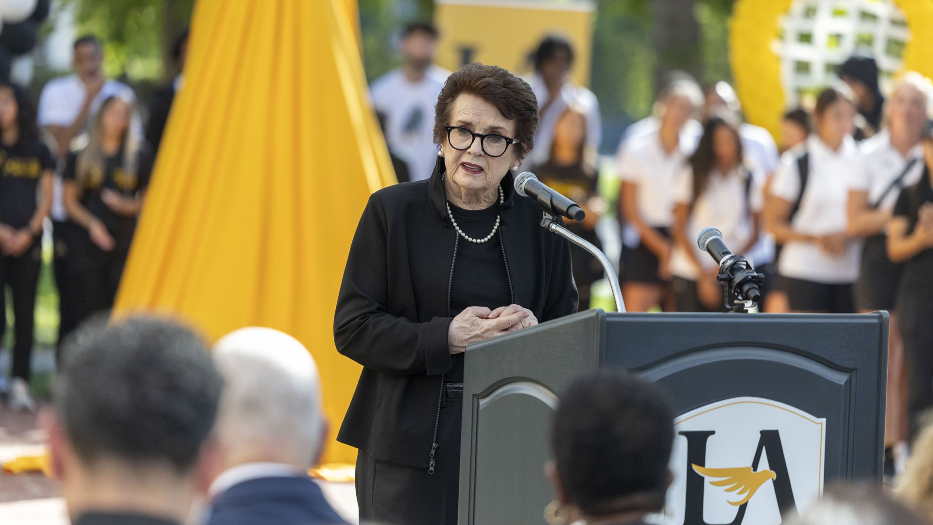 Speaker delivers remarks at a podium during an outdoor campus event, with attendees gathered in the background at Cal State LA. 