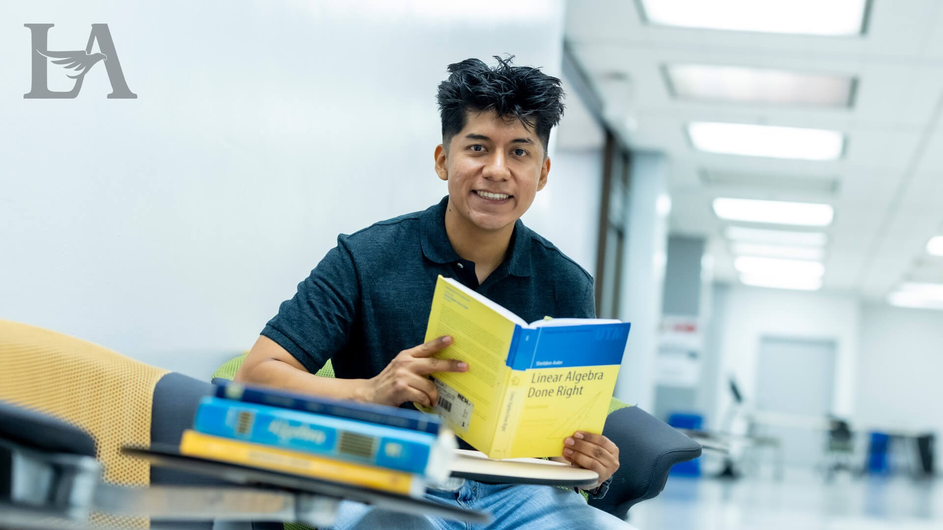 Student sits in a campus study area holding a linear algebra textbook, with additional math books in the foreground.