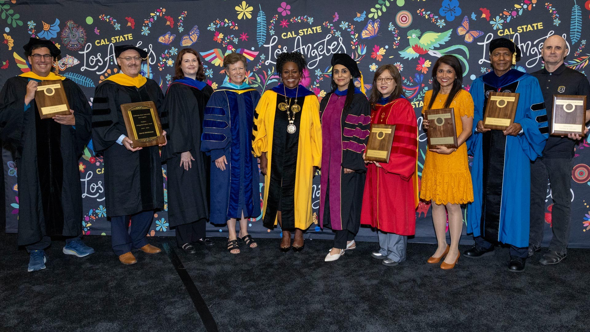Cal State LA faculty and staff pose with plaques during an investiture ceremony, standing in front of a vibrant backdrop featuring floral and bird illustrations.