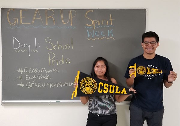 Two smiling students holding Cal State LA banners in front of chalkboard with the wordds GEAR UP written on it.