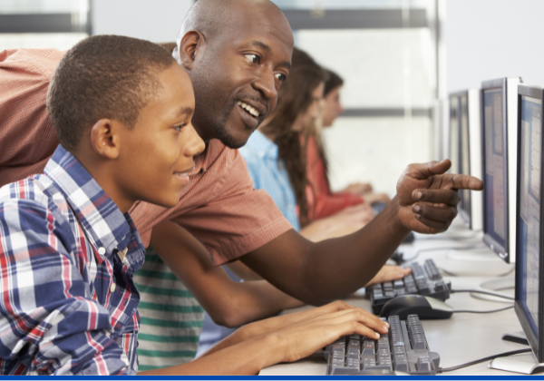 Male teacher leans over pointing to computer screen as young child looks on.
