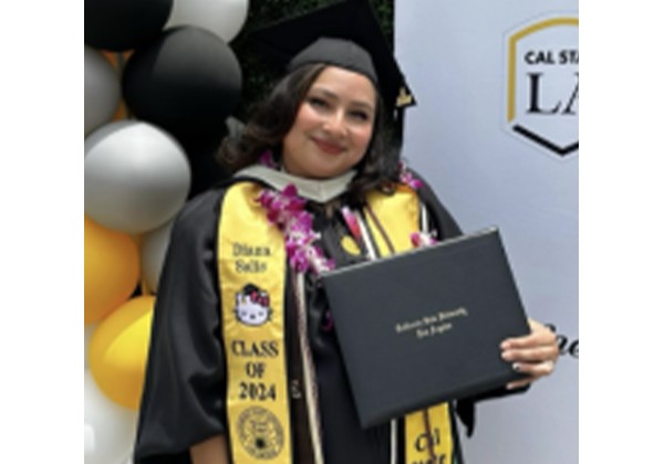 A smiling graduate wearing a cap and gown, leis, and decorated graduation stoles holds a diploma cover in front of a Cal State LA backdrop.