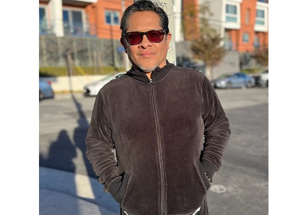 A man wearing red sunglasses and a dark zip-up jacket stands outdoors on a sunny street.