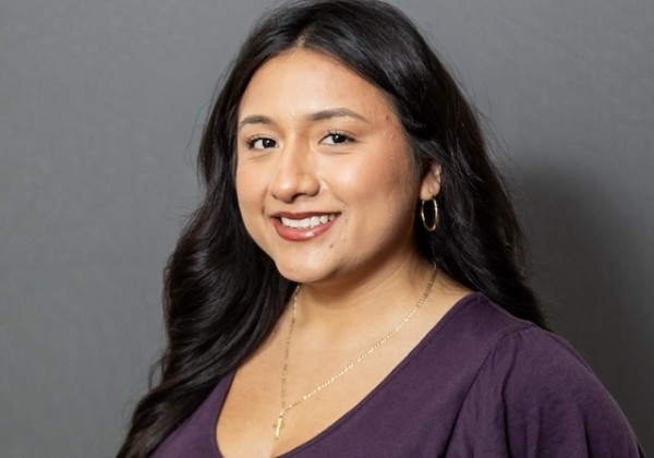 Guadalupe Garcia smiling with long dark wavy hair, wearing a purple top, gold hoop earrings, and a necklace against a gray background.
