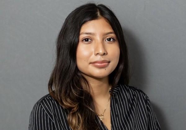 Itzel Gonzalez with long dark hair wearing a black pinstriped blouse and necklace, posed against a gray background.