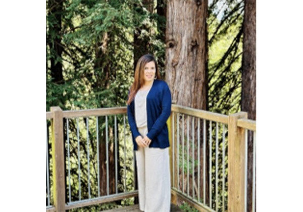 A woman wearing a blue cardigan and light-colored pants stands on a wooden deck surrounded by trees.