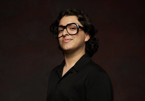 Portrait of Joey Rene, CREAR Futuros liaison, smiling slightly in a black shirt against a dark maroon background