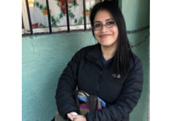 A woman wearing glasses and a black jacket smiles while seated indoors against a teal wall.
