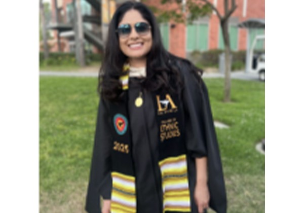 A smiling graduate wearing sunglasses, a black cap and gown, and decorated graduation stoles stands outdoors on a grassy campus walkway.
