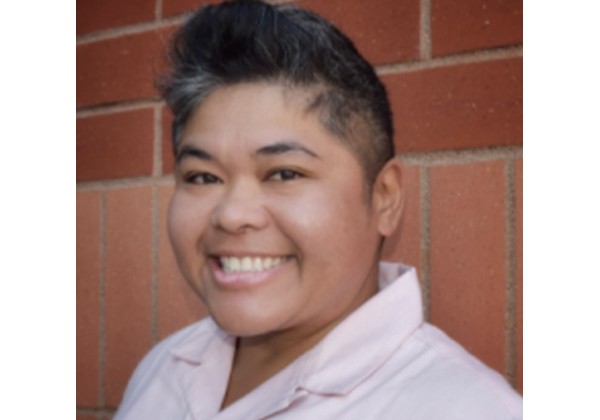 A smiling person wearing a light pink collared shirt poses in front of a brick wall.