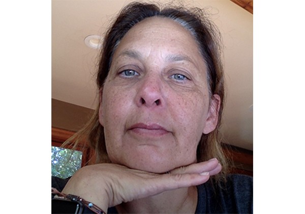 A woman rests her chin on her hand while looking toward the camera in a close-up indoor portrait.
