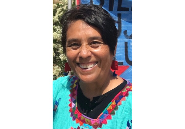 A smiling woman with short dark hair wears a turquoise embroidered top and red earrings while standing outdoors.