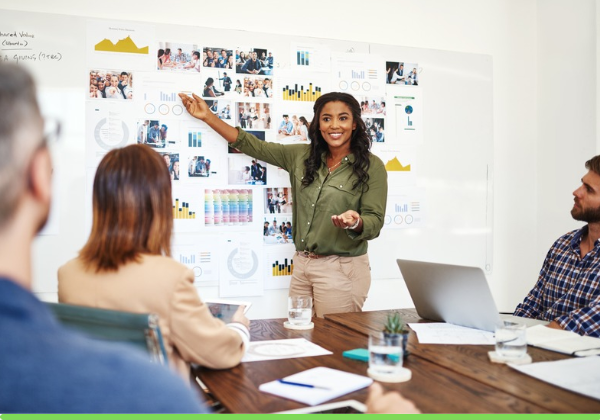Women stands in front of white board that has sticky notes attached making a presentation. Arms are open and she point to board as co-worker look on.