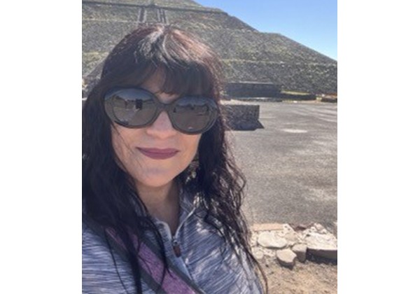 A woman wearing sunglasses takes a selfie outdoors in front of a large stepped pyramid under a clear blue sky.