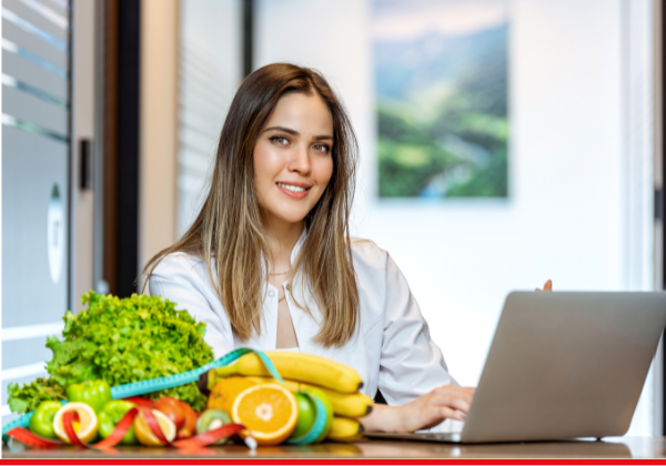 Female wearing white coat sits at desk with laptop and assortment of fruits and vegetables.