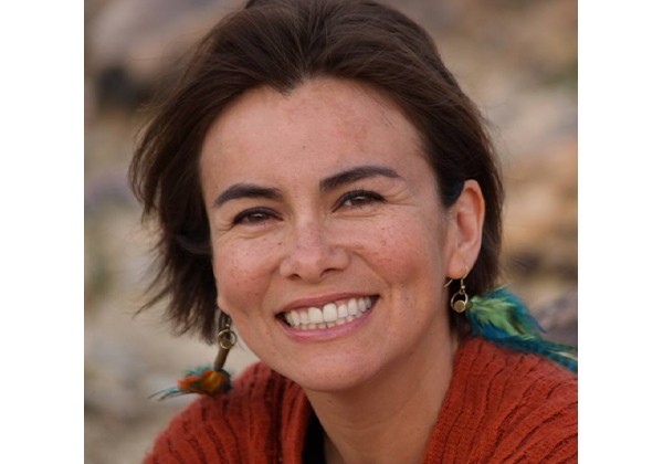A smiling woman with short brown hair wears feather earrings and a rust-colored sweater while posing outdoors against a softly blurred natural background.