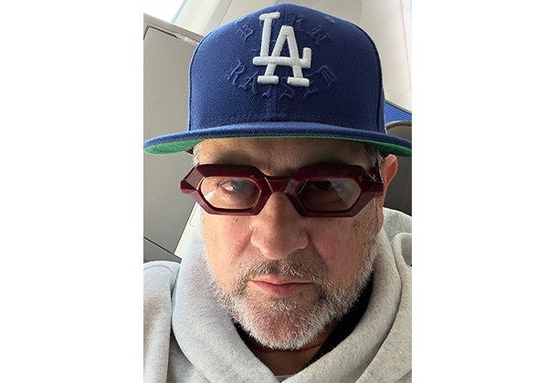 A man wearing red glasses, a gray hoodie, and a blue Los Angeles Dodgers cap looks toward the camera indoors.