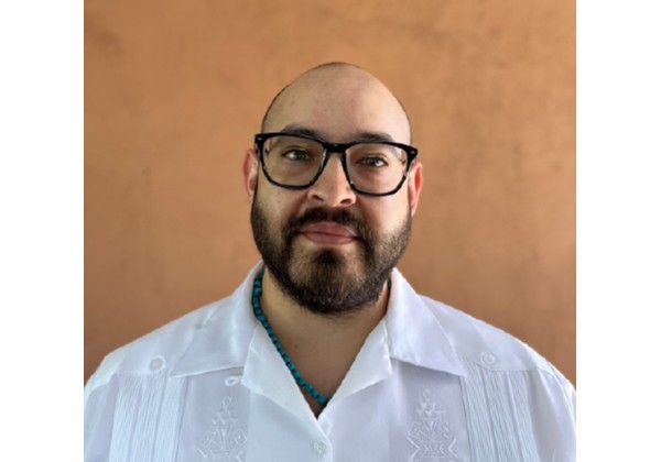 Pedro wearing glasses and a white shirt standing in front of a yellow wall