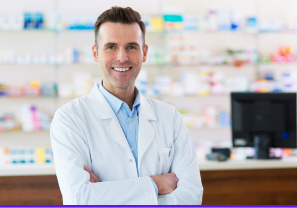 Male pharmacy tech stands with arms crossed. Shelves with medications are in the background.