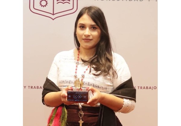 A woman wearing traditional-style clothing holds a decorative object while standing in front of a backdrop with a university-style logo and Spanish text.