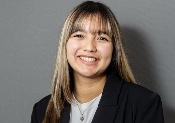Sonya Quintero Fernandez with long highlighted hair and bangs, wearing a black blazer over a white top against a gray background.