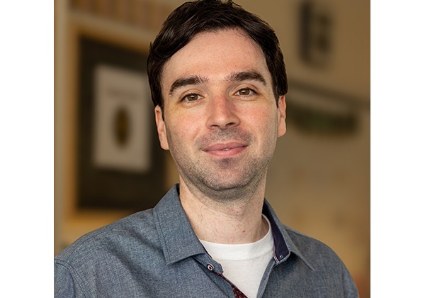 Tanner Leiderman wearing a grey dress shirt with a white t-shirt underneath standing inside an office
