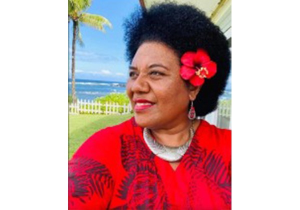 A woman wearing a red top and a red flower in her hair smiles outdoors near the ocean, with palm trees and a white fence in the background.