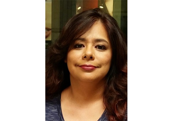 A woman with long wavy dark hair looks toward the camera in a close-up portrait taken indoors.