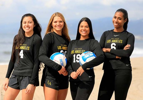 Four volleyball players standing on a beach, the two in the center are each holding a volleyball.