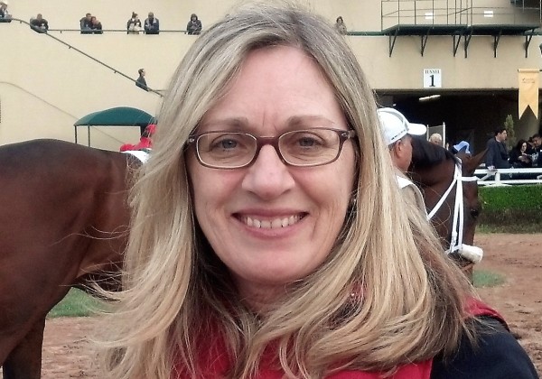 A woman at a horse track and smiling at the camera.