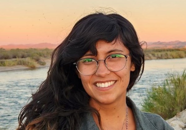 A woman with long hair standing in front of the a river smiling.