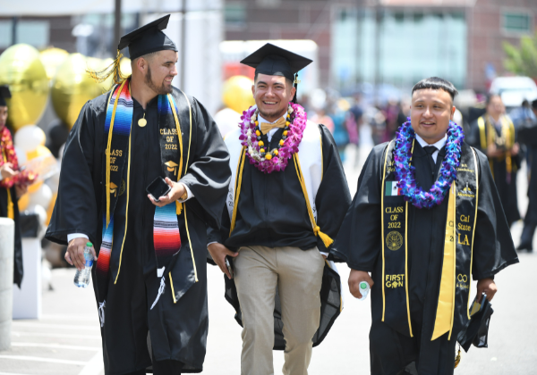 Three male students walking on campus wearing graduation regalia.