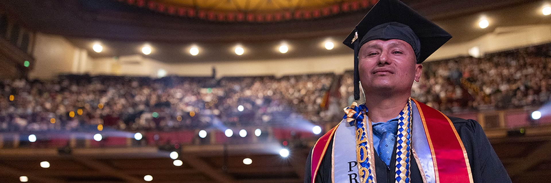 A student looking confident in graduation attire, behind him is a theatre full of people.
